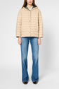 A woman stands wearing a beige doudoune capuche, blue flared jeans, and black shoes against a plain background. Her hands are by her sides, and her head is out of frame.