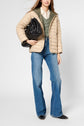 Woman in a beige doudoune fine, green sweater, white shirt, blue flared jeans, and black boots holds a large black bag against a plain background.