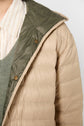 Close-up of a person wearing a beige doudoune réversible with an olive green lining, over a green sweater and a beige-and-white striped collared shirt. Only the upper torso is visible.