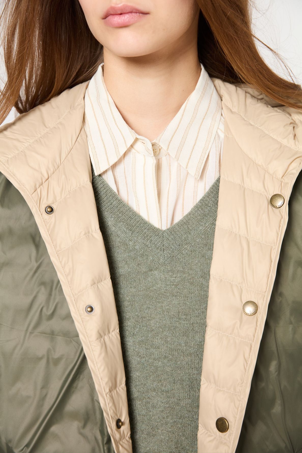 A person wearing a beige doudoune fine quilted jacket over a green V-neck sweater and a beige striped collared shirt. Only the lower half of their face and upper torso are visible.