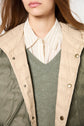 A person wearing a beige doudoune fine quilted jacket over a green V-neck sweater and a beige striped collared shirt. Only the lower half of their face and upper torso are visible.