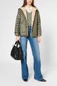 A woman wearing a green doudoune capuche, a green sweater, striped shirt, blue flared jeans, and black pointed shoes holds a large black handbag.