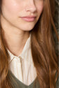 A close-up of a young woman with long, light brown hair, wearing a cream-colored collared shirt with thin stripes and a green sweater layered under a doudoune fine. Only her lower face and upper torso are visible.