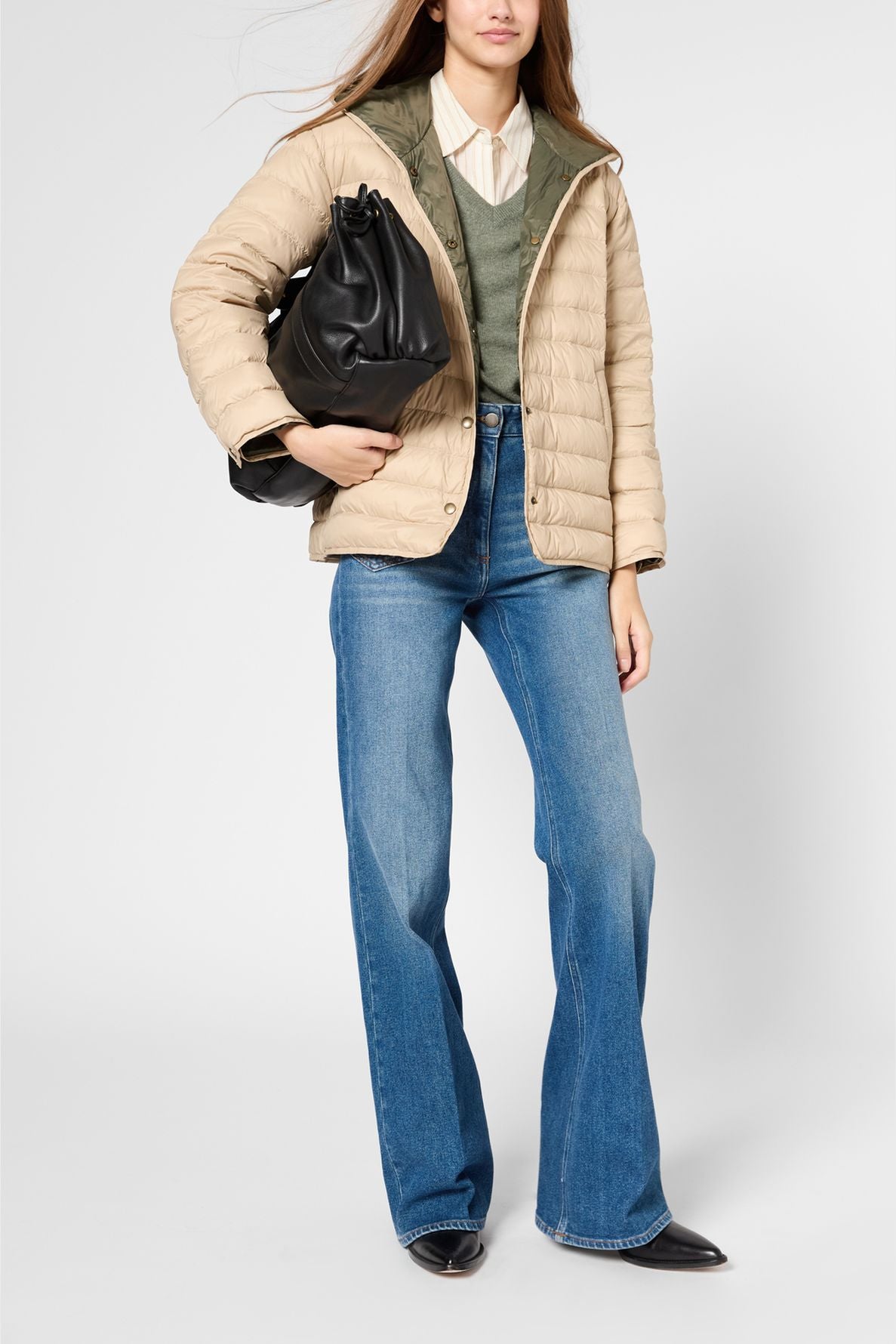 A person wearing a beige doudoune réversible, green sweater, white shirt, flared blue jeans, and black shoes holds a large black bag against a plain background.