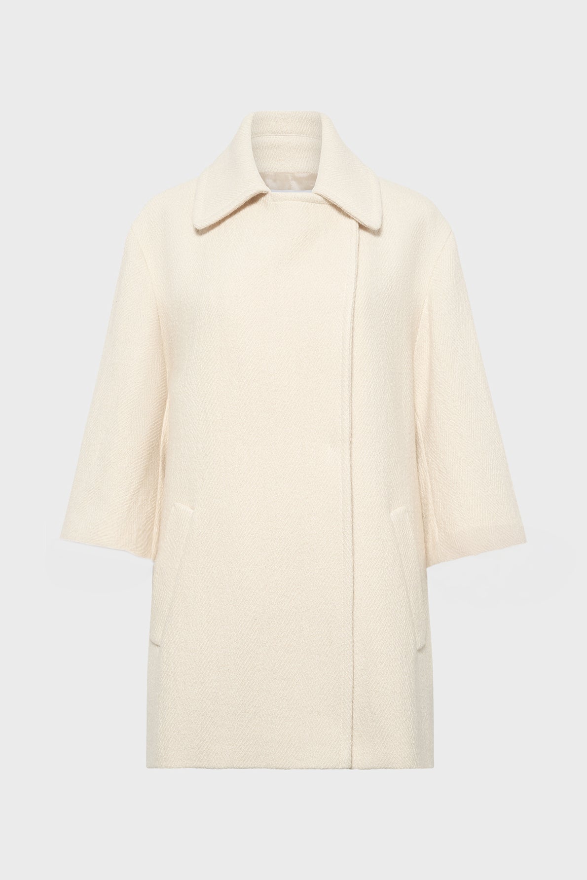 A cream-colored, textured manteau coton laine with a wide collar, three-quarter sleeves, and two front pockets, featuring a subtle motif chevrons and displayed on a plain light gray background.