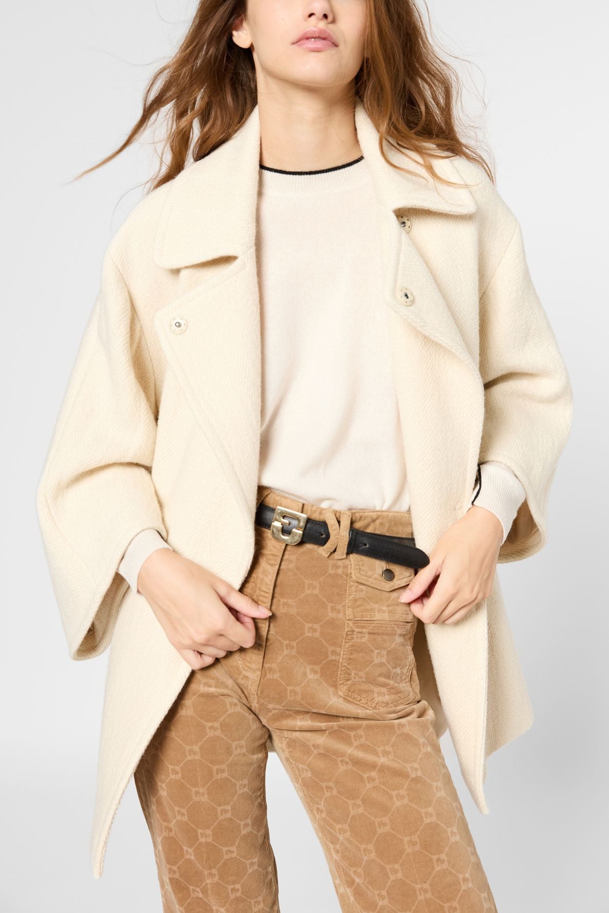A woman wears a manteau coton laine cream-colored coat over a light sweater and brown patterned pants with a black belt, posing with hands near her waist against a plain background.