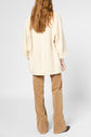 A person with long, wavy brown hair wears a cream-colored manteau coton laine and tan patterned pants adorned with motif chevrons, standing with their back facing the camera against a plain background.