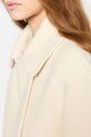 Close-up of a person with long brown hair wearing a cream-colored manteau coton laine with a large collar. The image highlights the coat’s texture, featuring a subtle motif chevrons, and shows part of the person’s face and neck.