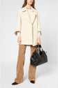 Woman wearing a cream manteau coton laine, beige patterned pants with motif chevrons, and black pointed shoes, holding a large black handbag with gold hardware, standing against a plain light background. Her hair is loose and she is partially out of frame.
