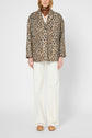 A person wearing a manteau léopard chic, white wide-leg pants, a brown patterned scarf, and beige platform sandals stands against a plain light background.