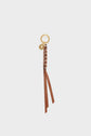 Gold keyring with a small round charm en cuir and a braided brown leather strap, reminiscent of a classic scoubidou, ending in two long leather tassels—an elegant accessoire sac displayed on a plain white background.