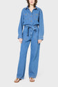 A person wearing a blue denim femme jumpsuit with a tied waist, long sleeves, front pockets, and brown sandals stands facing forward against a plain light background.