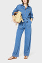 Person standing in a blue denim femme jumpsuit with long sleeves and wide legs, featuring a ceinture amovible, holding a woven beige handbag, and wearing brown flat sandals. The background is plain white.