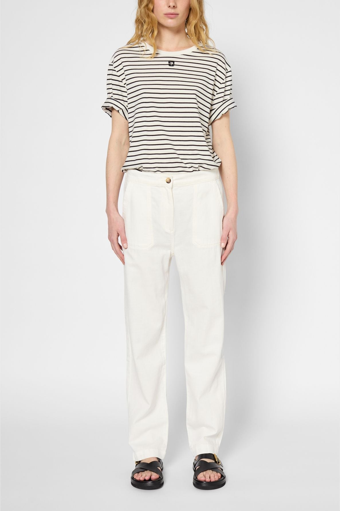 A person stands facing forward, wearing a black and white striped t-shirt, loose pantalon blanc in coton lyocell lin, and black sandals against a plain light background. The person’s arms are relaxed at their sides.