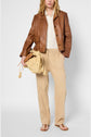A person wearing a brown leather jacket, cream shirt, and a chic pantalon camel in coton lyocell lin with flip-flops holds a textured woven bag with tassels. The background is plain white.