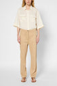 Person standing against a plain background, wearing a cream short-sleeve button-up shirt with chest pockets, light beige pantalon droit crafted from a coton lyocell lin blend, and brown sandals. The face is partially visible.
