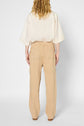 A person stands facing away, wearing a loose cream-colored shirt and a pantalon droit in a high-waisted beige shade with back pockets, crafted from coton lyocell lin. Their wavy blonde hair rests on their shoulders. The background is plain and light.