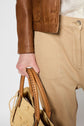 A person wearing a pantalon droit in camel—a stylish blend of coton, lyocell, and lin—paired with a brown leather jacket holds a woven straw bag with stitched leather handles.