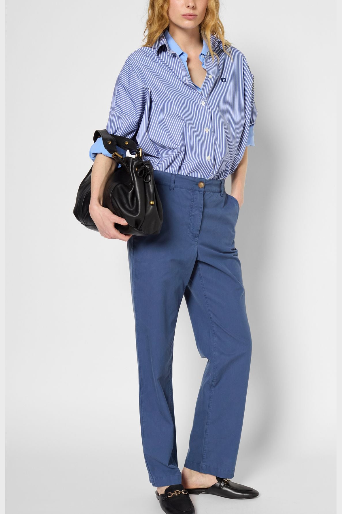 Person wearing a blue striped button-up shirt tucked into a pantalon large taille haute in blue, holding a large black handbag and wearing black loafers, standing with one hand in their pocket against a plain background.