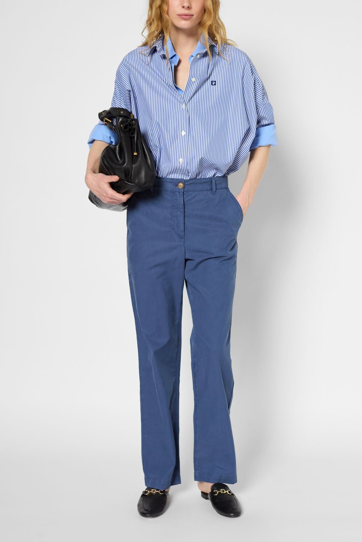 Person in a blue striped button-up shirt with rolled sleeves, loose taille haute blue trousers in lin et lyocell, and black loafers, holding a black bag under one arm, standing against a plain light background.