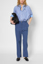 Person in a blue striped button-up shirt with rolled sleeves, loose taille haute blue trousers in lin et lyocell, and black loafers, holding a black bag under one arm, standing against a plain light background.