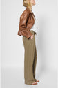 Woman in a brown leather jacket, white top, and loose-fitting olive pants stands in profile against a plain light background, wearing sandals.