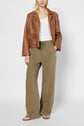 Woman wearing a brown leather jacket, white top, loose olive green pants, and tan sandals, standing against a light gray background. Her hands are in her pockets and her hair is loose.
