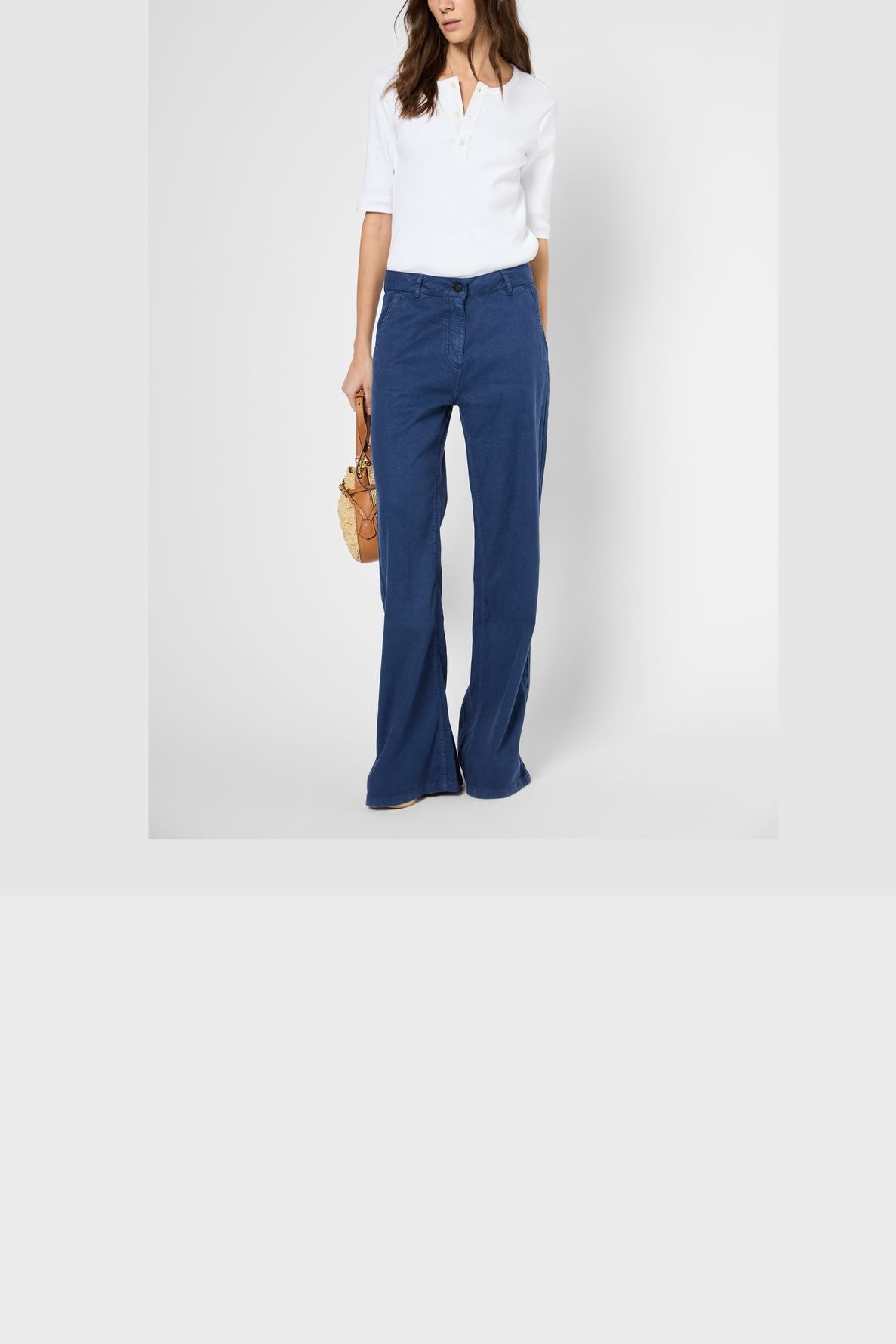 Woman wearing a white short-sleeve blouse and blue flared pants, holding a small beige handbag, standing against a plain white background. Her head is partially out of frame.