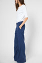 Woman standing sideways, wearing a white 3/4 sleeve top and high-waisted, wide-leg blue pants with hands in pockets. Background is plain and white.