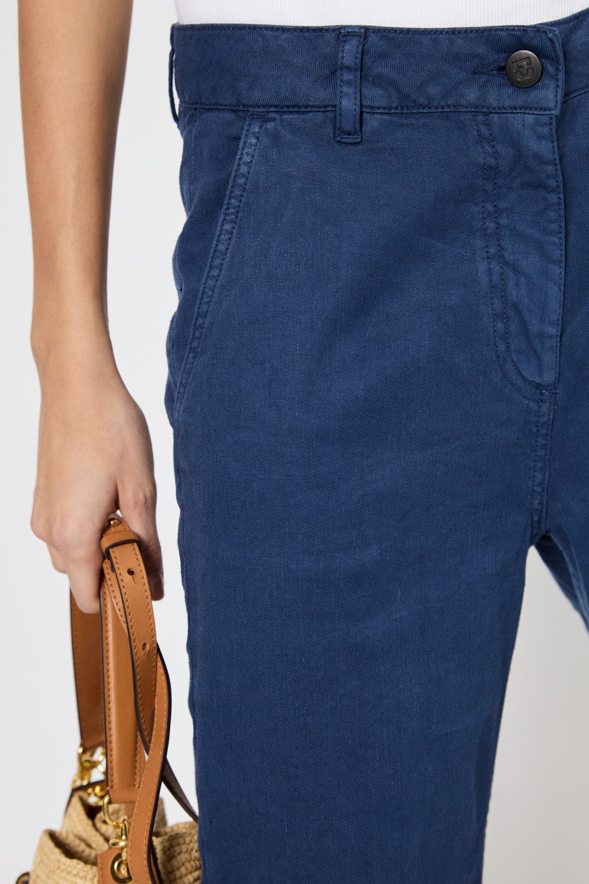 A person wearing blue pants and a white top holds a tan handbag by its handles. The image is cropped to show only the lower torso and part of one arm.