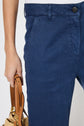 A person wearing blue pants and a white top holds a tan handbag by its handles. The image is cropped to show only the lower torso and part of one arm.