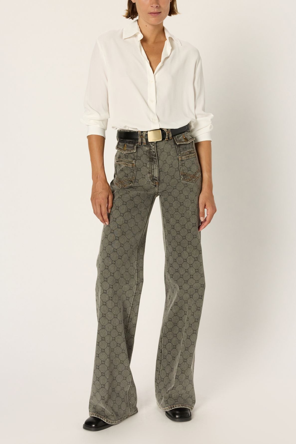 A person wearing a white button-up shirt, gray flared jeans à motif géométrique taille haute with a black belt, and black shoes stands against a plain white background.