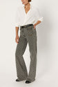 Person wearing a white button-up shirt tucked into high-waisted, wide-leg gray patterned jeans featuring a motif géométrique, with a black belt and black shoes, standing against a plain white background.