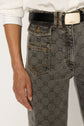 A person wearing patterned gray jeans with a motif géométrique and front pockets, a black belt with a gold buckle, and a white shirt. Only the lower torso and hand are visible.