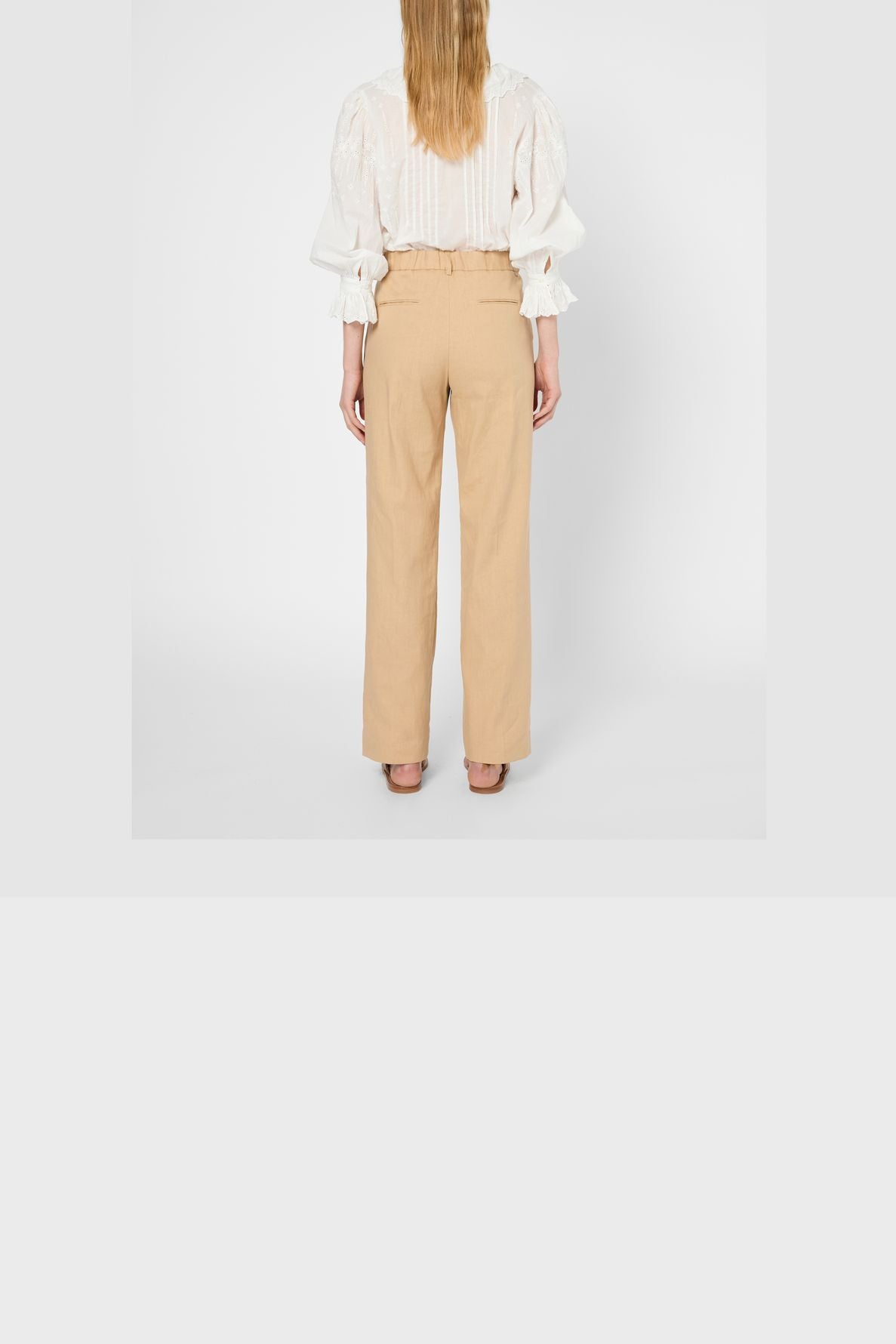 A person with long blonde hair, seen from the back, wears a white blouse with puffed sleeves and a high-waisted pantalon large femme in beige linen-cotton, paired with brown shoes, standing against a plain white background.