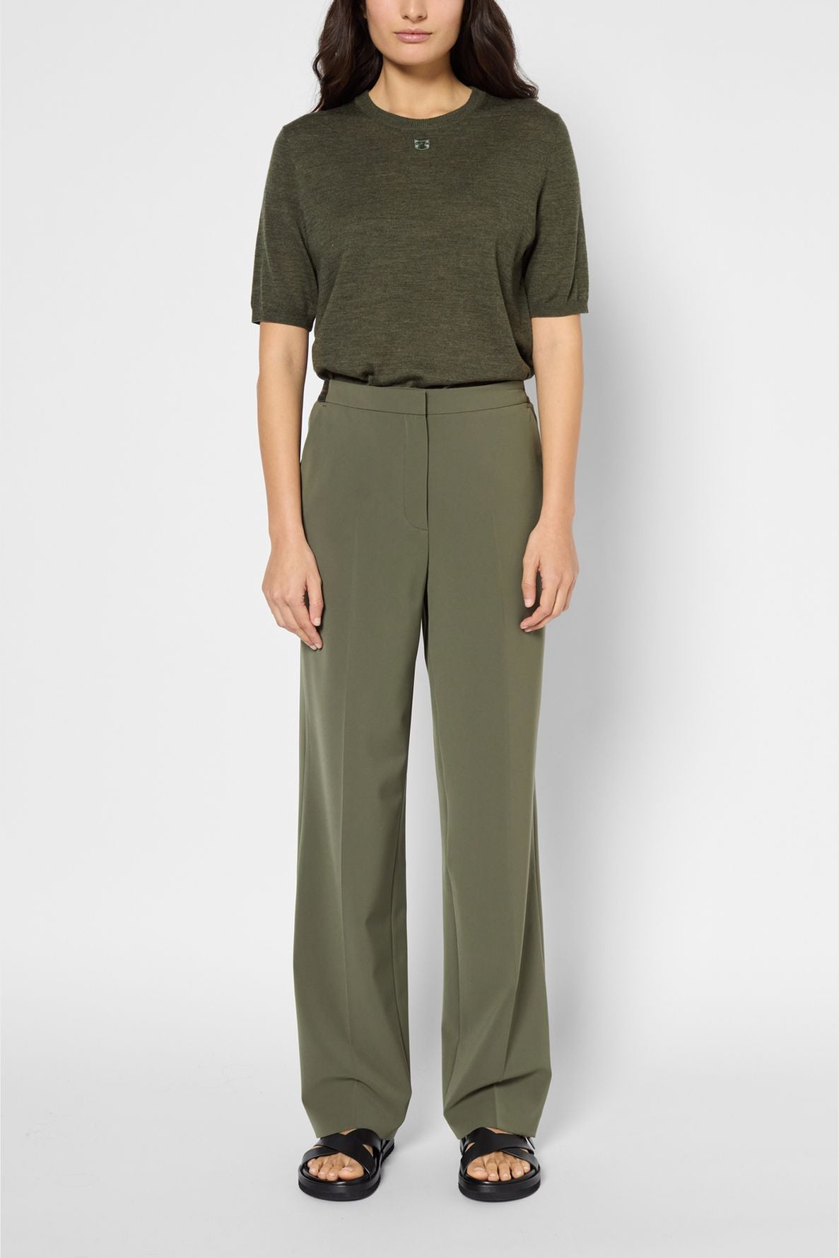 A woman stands against a plain background wearing an olive green short-sleeve top and a pantalon large femme in microfibre kaki, paired with black sandals. Her hair is loose and she has a neutral expression.