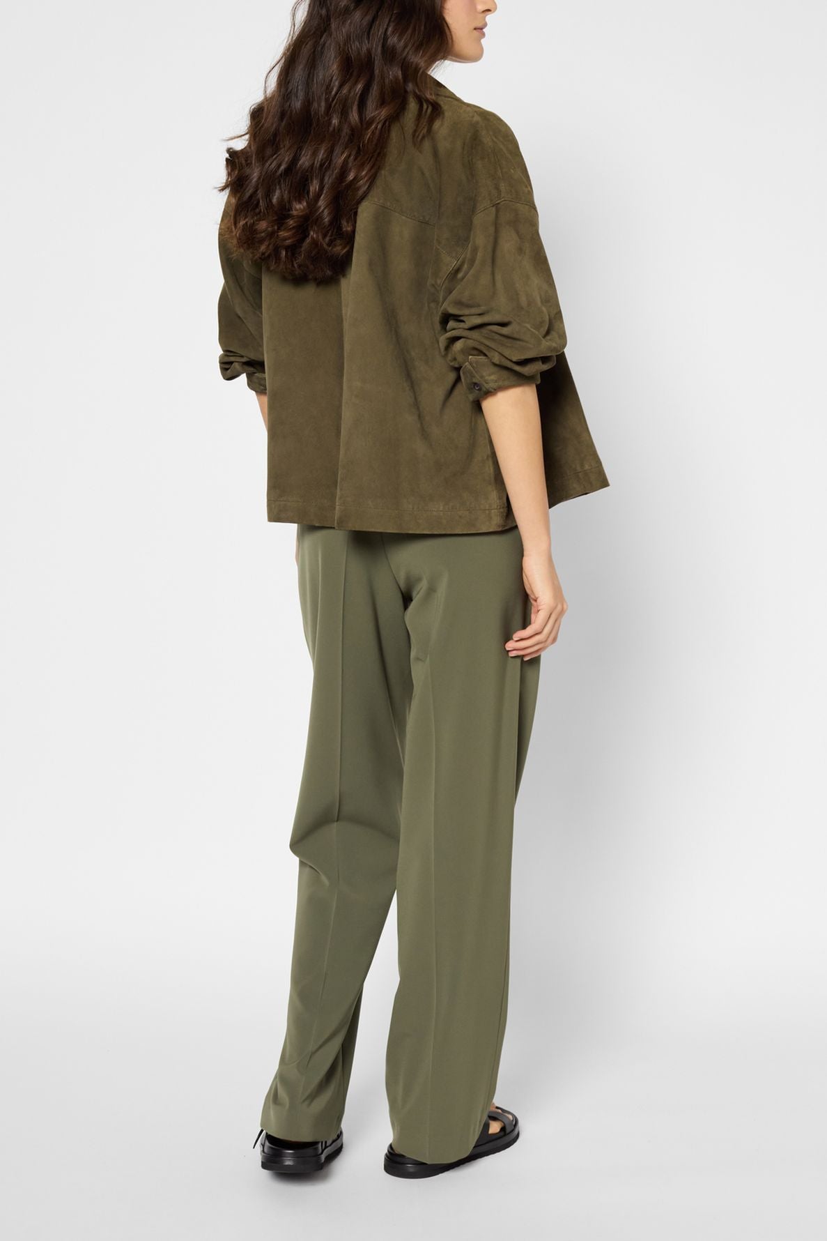 A woman is standing with her back to the camera, wearing an olive green jacket and a pantalon fluide kaki paired with black shoes. Her long, wavy brown hair is down. The background is plain and light gray.