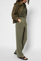 A woman wearing an olive green microfibre kaki jacket, matching pantalon fluide kaki, and black sandals stands with her hands in her pockets against a plain white background. Her face is partially out of frame.