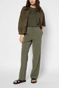 A person wearing an olive green top, a pantalon large femme in microfibre kaki, a brown jacket, and black sandals stands against a plain background with one hand in their pocket. Their face is partially out of frame.