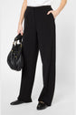 Person wearing a chic pantalon large in black microfibre paired with sleek black loafers featuring gold detail, holding a black leather handbag in one hand, standing against a plain white background. Only the lower half of the body is visible—evoking understated elegance in a modern tailleur femme aesthetic.