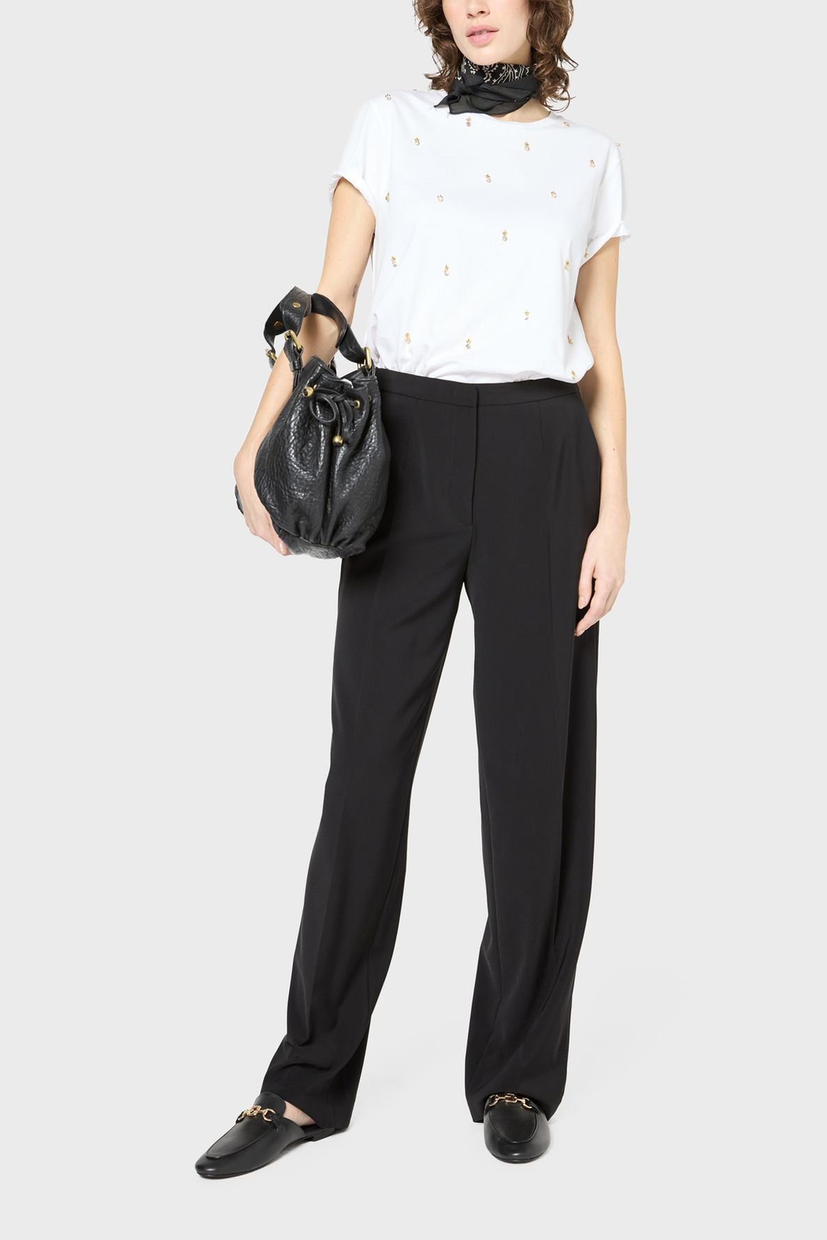 A person wearing a white t-shirt with small patterns, black pantalon large crafted from soft microfibre, black loafers, a black scarf, and carrying a large black handbag stands against a plain background.