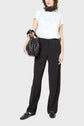 A person wearing a white t-shirt with small patterns, black pantalon large crafted from soft microfibre, black loafers, a black scarf, and carrying a large black handbag stands against a plain background.
