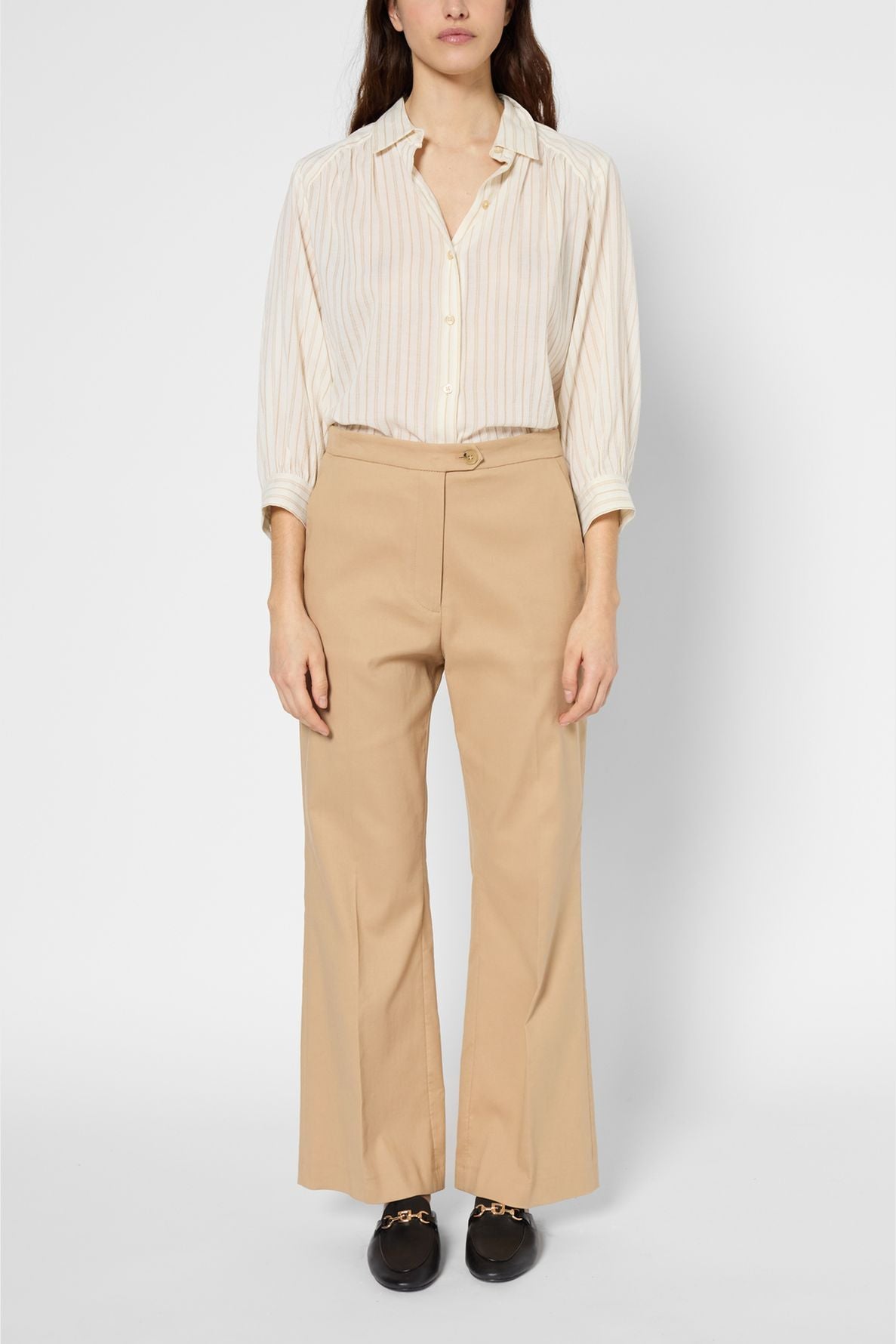 Woman stands facing forward, wearing a cream striped button-up shirt tucked into tan pantalon évasé and black loafers. Her arms rest by her sides, and her long hair is down. The background is plain and white.