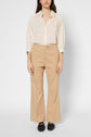 Woman stands facing forward, wearing a cream striped button-up shirt tucked into tan pantalon évasé and black loafers. Her arms rest by her sides, and her long hair is down. The background is plain and white.