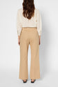 A woman stands facing away, wearing a cream striped blouse and tan high-waisted, wide-leg pantalon évasé. Her long brown hair is loose, and she is wearing black shoes. The background is plain white.