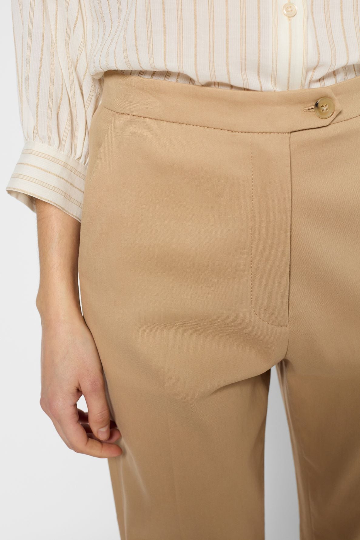 A person wearing a pantalon droit in tan dress pants with a button and zipper, and a cream, striped button-up shirt with rolled sleeves. Only the lower torso and one hand are visible against a neutral background.