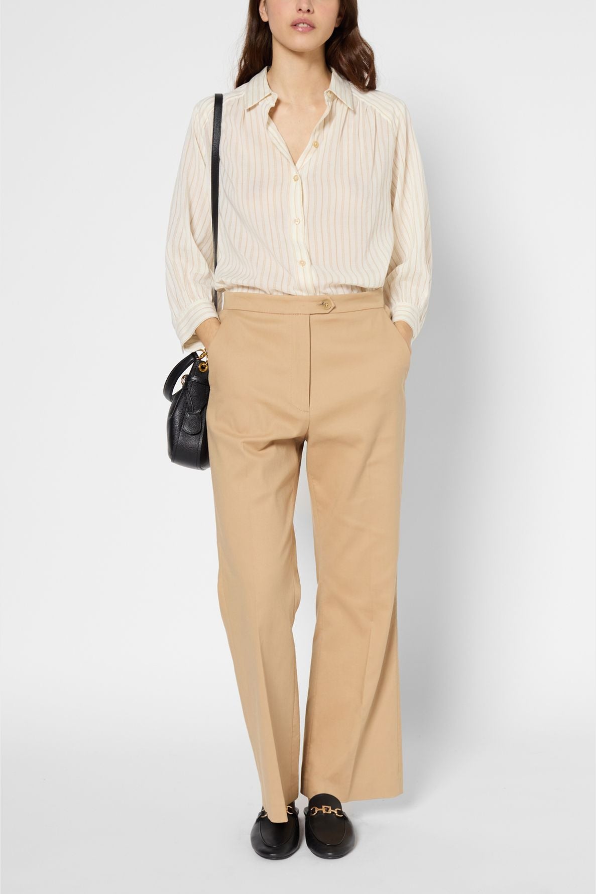 Woman wearing a beige striped button-up shirt, tan high-waisted pantalon évasé, black loafers with gold detail, and a black shoulder bag, standing with hands in pockets against a plain background.