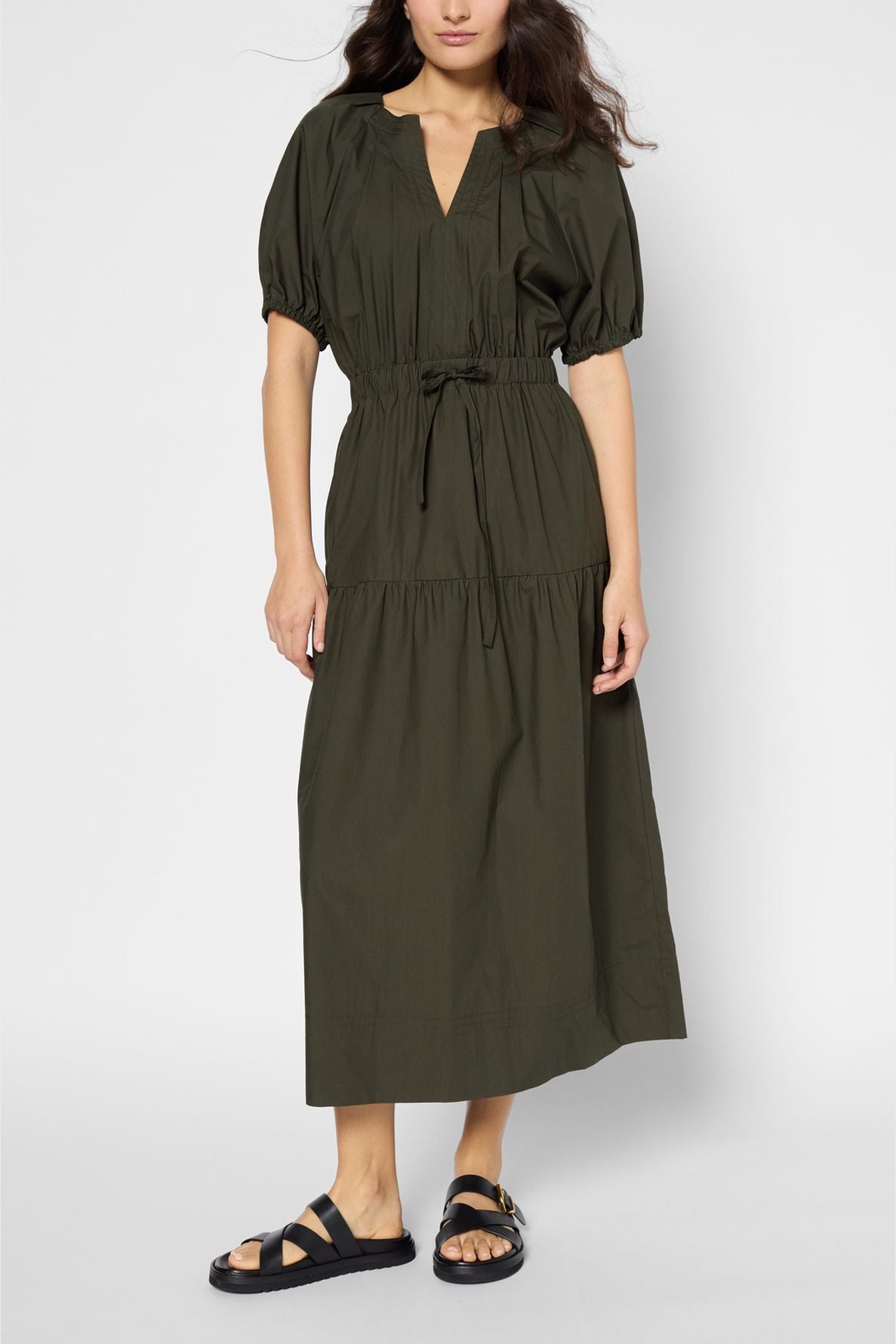 A woman wears a dark green popeline kaki, short-sleeve, midi-length dress with a gathered waist and tiered skirt. She pairs this élégante coton robe longue with black strappy sandals. The background is plain white.
