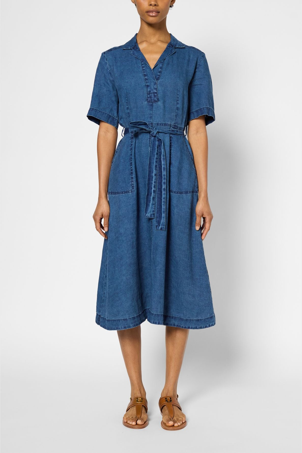 A person wears a knee-length, short-sleeved denim dress with a collar, front buttons, a waist tie, and front pockets, paired with brown sandals.