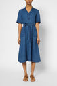 A person wears a knee-length, short-sleeved denim dress with a collar, front buttons, a waist tie, and front pockets, paired with brown sandals.